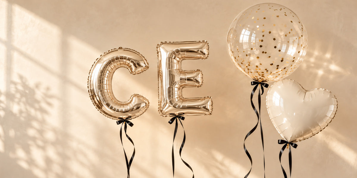 Champagne-gold C and E letter balloons beside a clear gold-confetti balloon and an ivory heart balloon, all with black ribbons