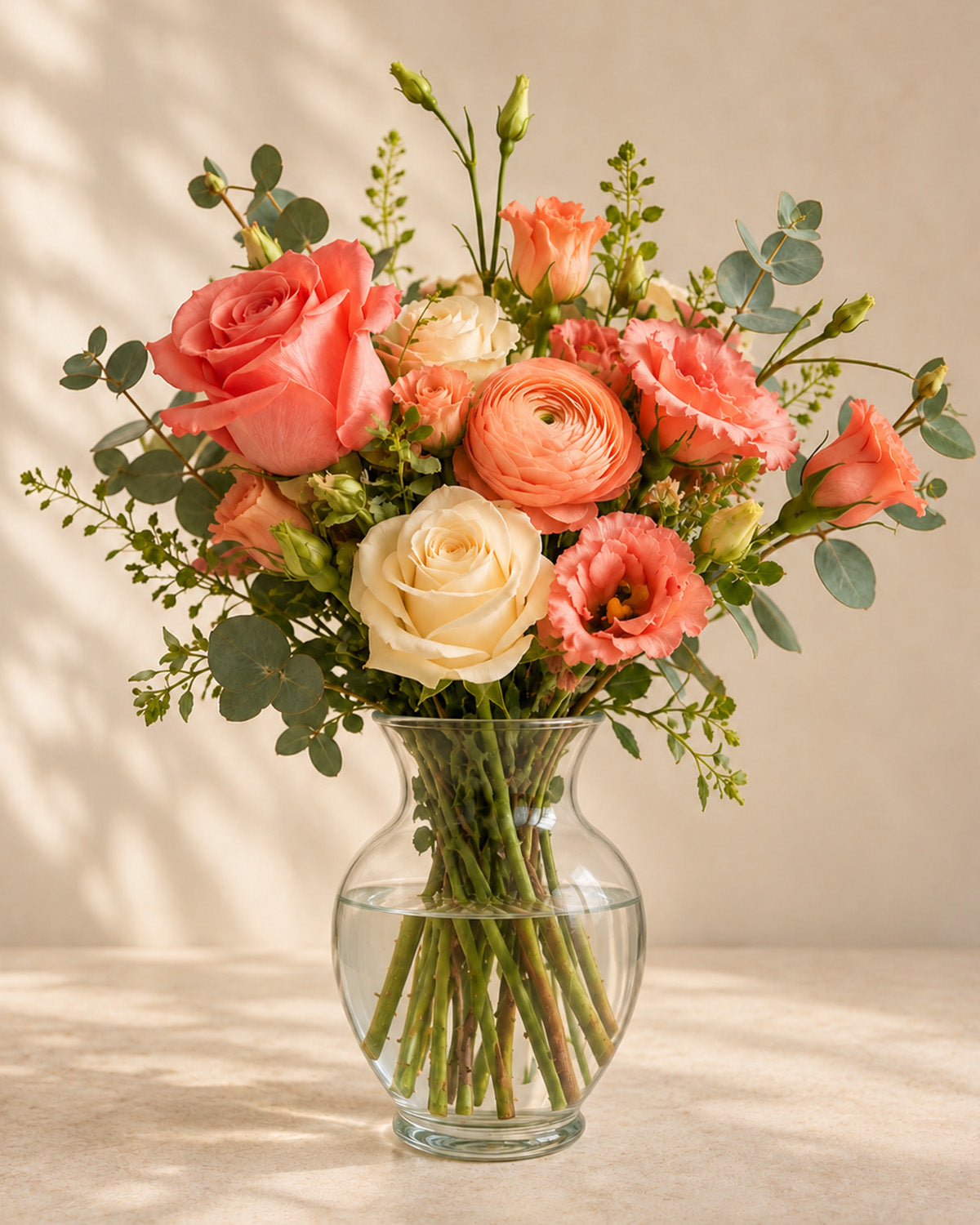 Coral and cream roses, raunculus, and eucalyptus arranged in a clear glass vase