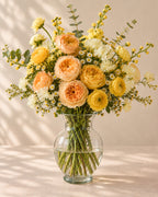 Champagne Morning bouquet with peach garden roses, yellow ranunculus, white flowers, and eucalyptus arranged in a clear glass vase
