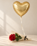 Single live red rose wrapped in cream paper and tied with a burgundy ribbon beneath a gold heart-shaped foil balloon