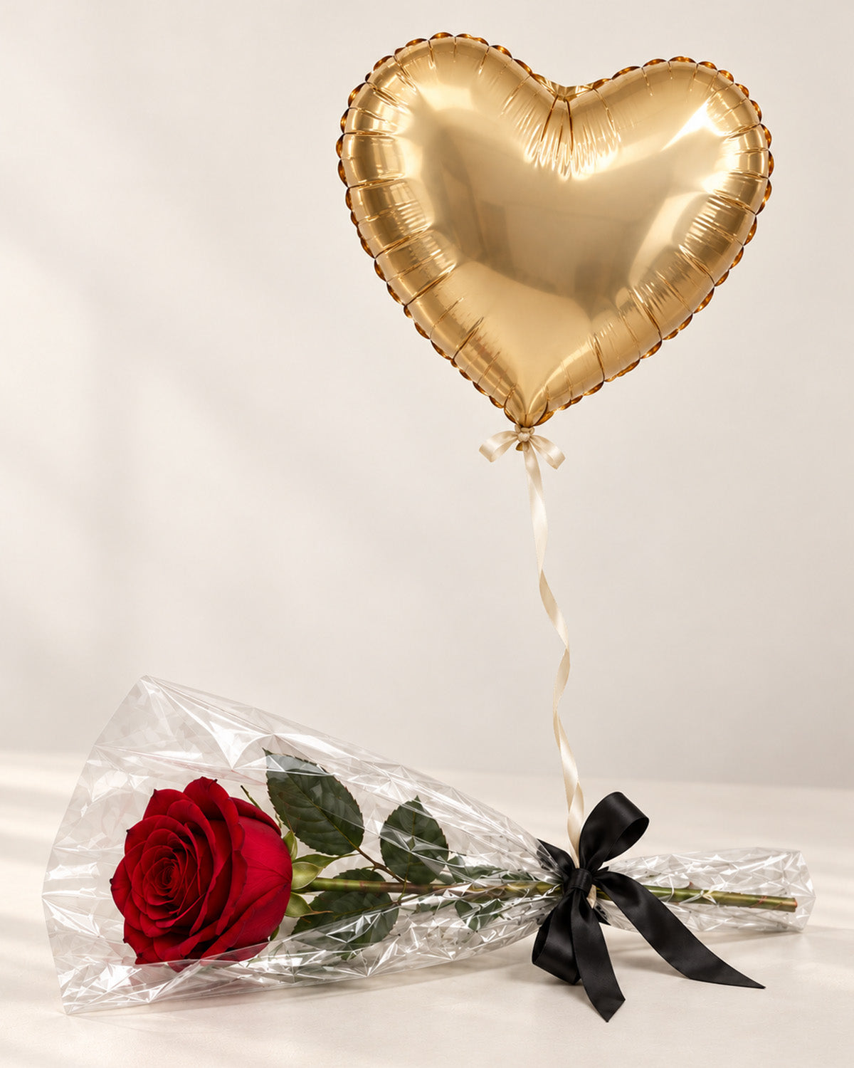 Single preserved red rose wrapped in clear cellophane and tied with a black ribbon beneath a gold heart-shaped foil balloon