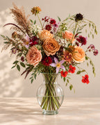 Vintage Ember bouquet with caramel roses, burgundy flowers, soft peach blooms and airy greenery in a clear glass vase